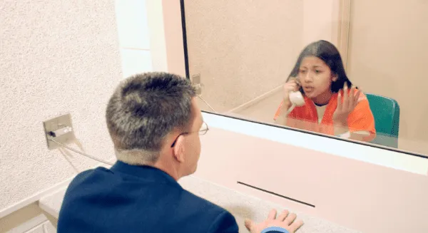 A Krayemandco criminal lawyer in a blue suit speaking through a glass partition to a client in an orange jumpsuit during a prison legal visit.