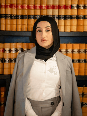 Professional portrait of a female Krayemandco criminal lawyer wearing a hijab, white shirt, and grey blazer, in front of a law library.
