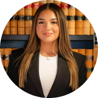 Professional portrait of a female Krayemandco criminal lawyer with long brown hair, wearing a black blazer, standing in front of a law library.