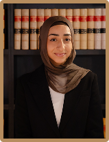 Professional portrait of a Krayem & Co female team member wearing a black blazer and brown hijab, standing in front of a law library bookshelf.