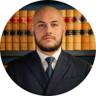 Professional portrait of a male Krayemandco criminal lawyer with a beard and shaved head, wearing a dark navy suit and patterned tie, in front of a law library.