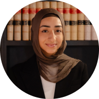 Circular portrait of a Krayemandco criminal lawyer in a black blazer and brown hijab, standing in front of legal library shelves. Representing our Criminal defence firm Sydney CBD office.