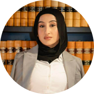 Professional portrait of a female Krayemandco criminal lawyer wearing a black hijab and light grey blazer, standing in front of a law library.