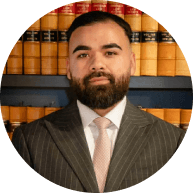 Professional portrait of a male Krayemandco criminal lawyer with a beard, wearing a pinstripe suit and light pink tie, in front of legal law reports.