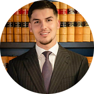 Professional portrait of a male Krayemandco criminal lawyer with a beard, wearing a dark suit and patterned tie, in front of a law library.