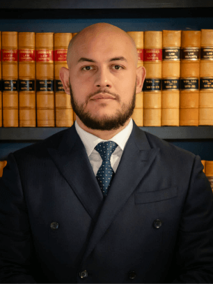 Professional portrait of a male Krayemandco criminal lawyer with a beard and shaved head, wearing a dark navy suit and patterned tie, standing in front of legal law reports.