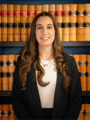 Professional portrait of a female Krayemandco criminal lawyer with long brown hair, wearing a navy blazer and white top, standing in front of a law library.