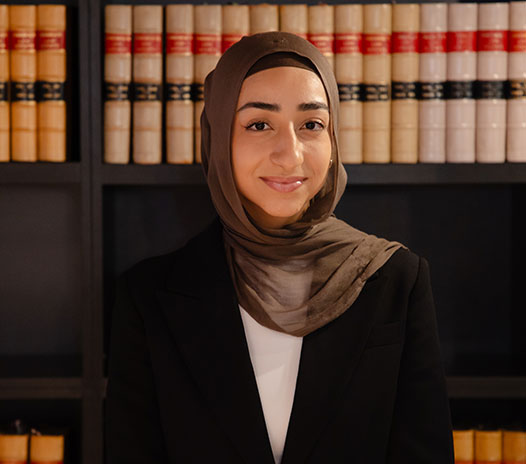 Professional portrait of a Krayemandco criminal lawyer wearing a black blazer and brown hijab standing in front of legal library shelves at our Criminal defence firm Sydney CBD.
